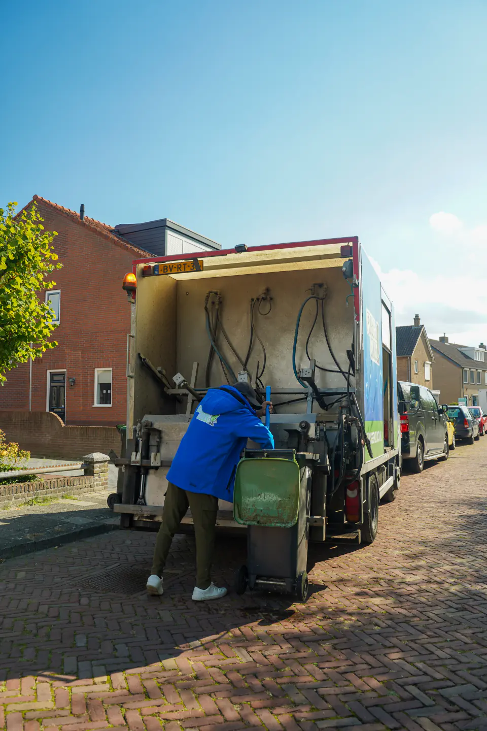 Medewerker van Frisse Kliko in regenjas zet een groene container klaar bij de reinigingswagen.
